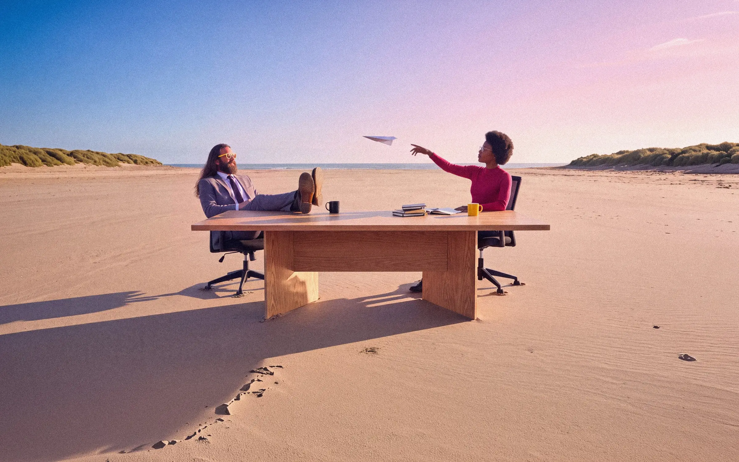 Twee mensen aan een houten tafel midden op een verlaten strand, een papieren vliegtuigje in de lucht